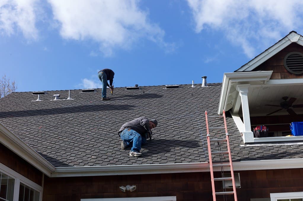 Two roofers working on a grey shingle roof under a clear blue sky