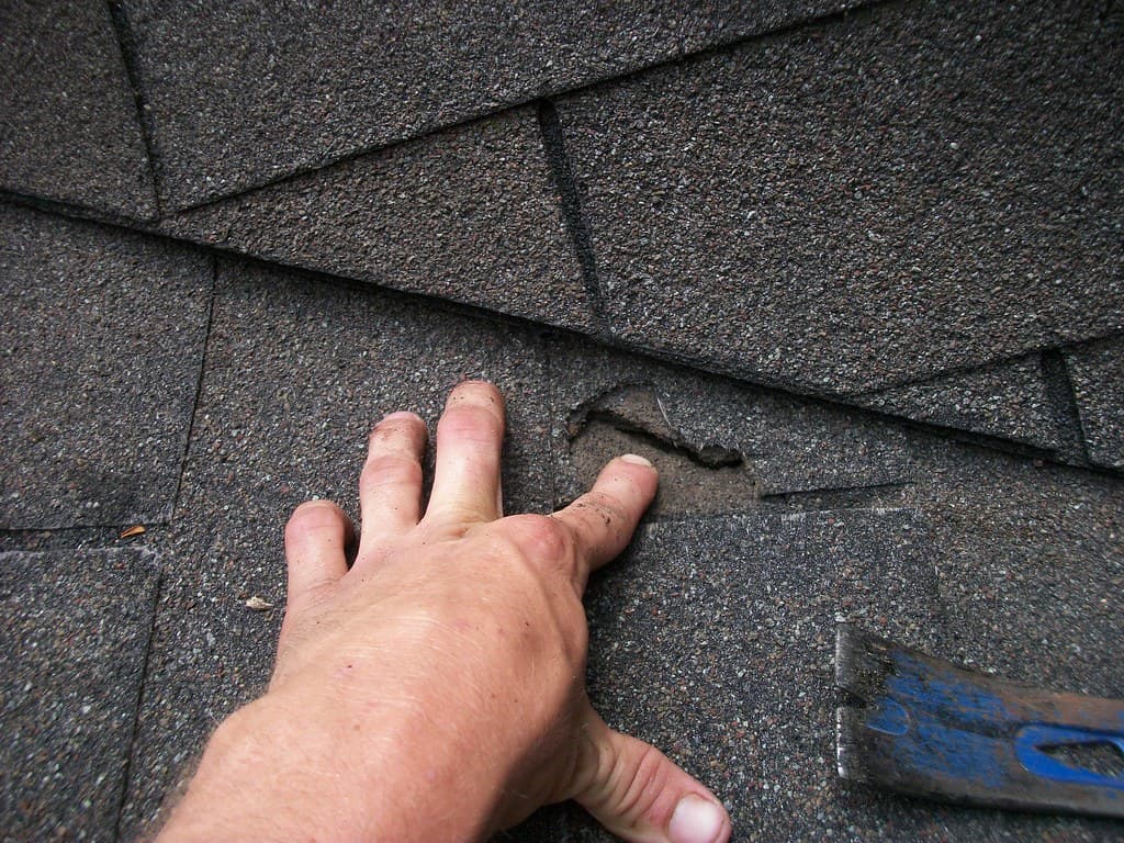 A tradesperson's hand inspecting a damaged roof shingle beside a roofing hammer